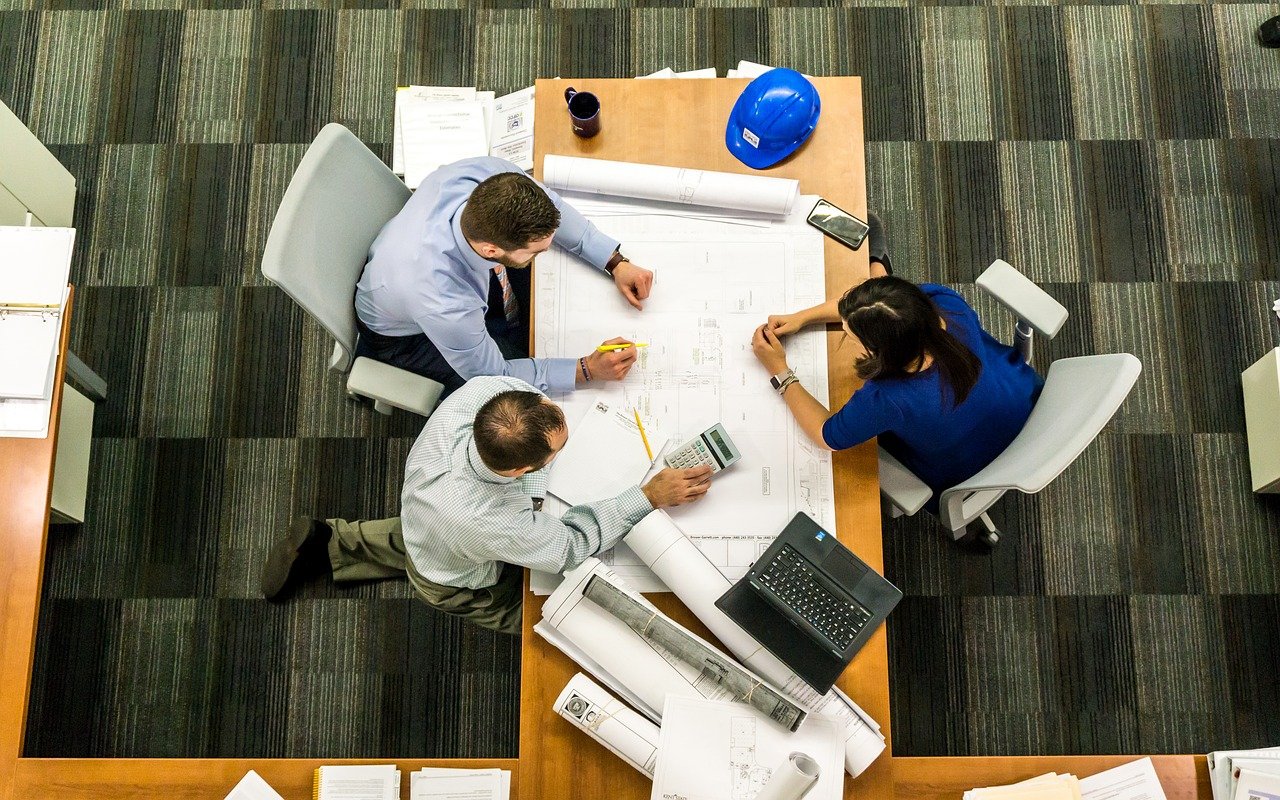 Photo from above a table, showing three people sitting around a large architect's drawing.